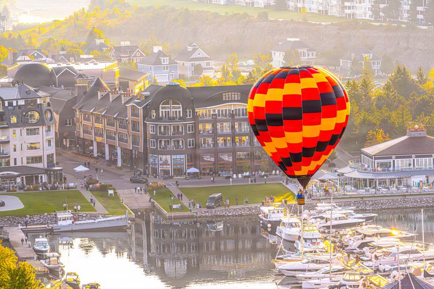 A red, black and yellow checkered hot air balloon hanging just above the Bay Harbor marina and the Main Street buildings in golden early light