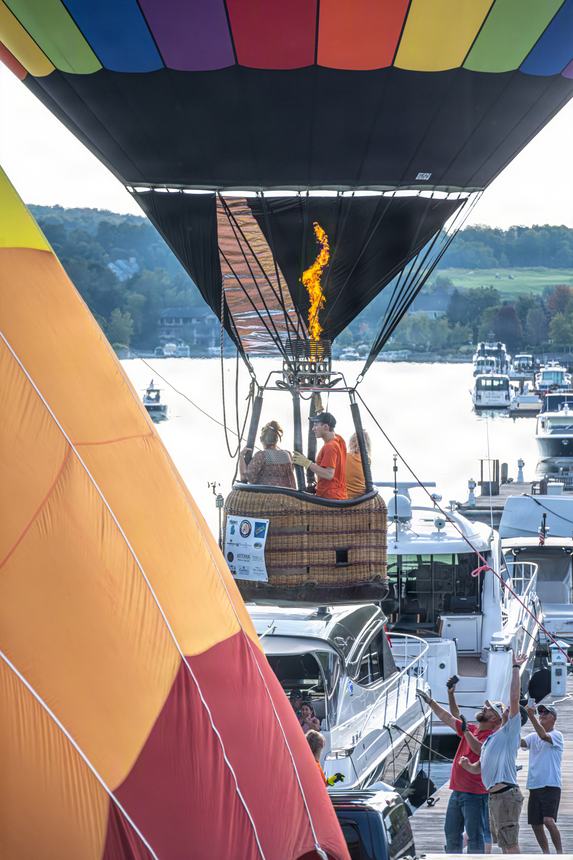 A wicker balloon basket carrying two passengers lifts past the marina, burner lit, spectators below raising their arms