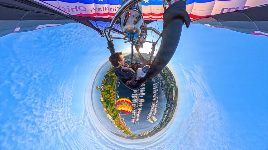 A 360-degree little-planet photograph from the basket with a second orange and yellow balloon flying alongside above the marina