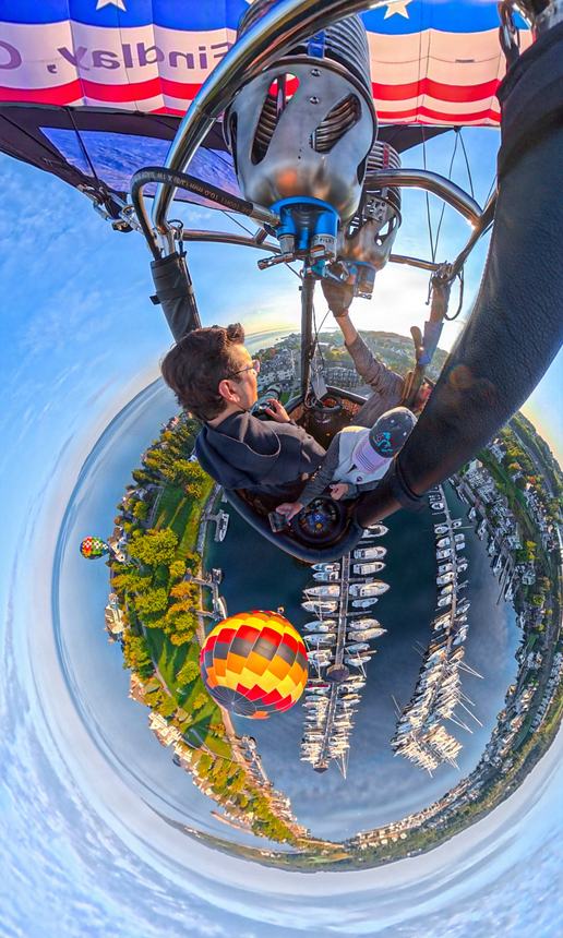 A 360-degree little-planet photograph of a passenger in the balloon basket with the Bay Harbor marina curved into a circle beneath and an orange balloon alongside