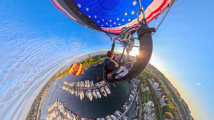 A 360-degree little-planet photograph from the balloon basket, the marina and village curled into a sphere below, sun bursting past the envelope