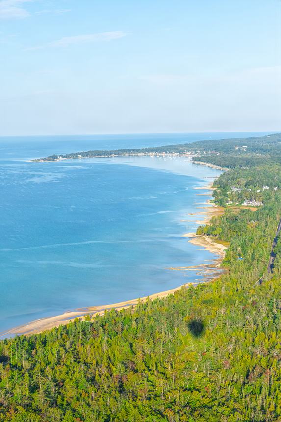 Aerial of the wooded Little Traverse Bay shoreline curving toward town, pale beach and clear blue water