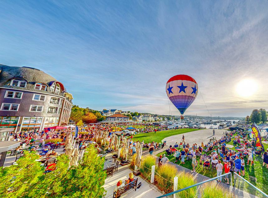 Wide view of the Bay Harbor Main Street buildings and the lawn full of spectators with a star-patterned balloon aloft over the marina in bright evening sun