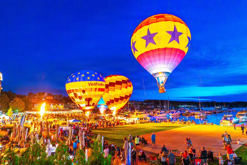 Three hot air balloons glowing from within at blue hour on the lawn beside the Bay Harbor marina, a crowd watching and boats lit along the docks