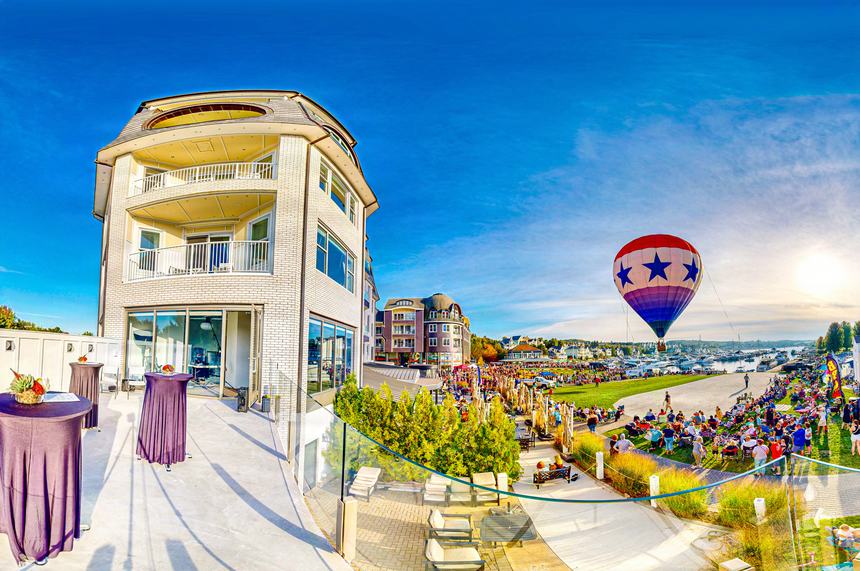 A red, white and blue star-patterned hot air balloon aloft over a crowd gathered on the Bay Harbor lawn at sunset, seen from a terrace above Main Street