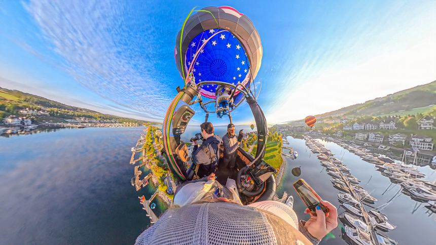 Wide 360-degree view from a balloon basket: the blue star-spangled envelope overhead, passengers below, and the Bay Harbor marina wrapping the horizon